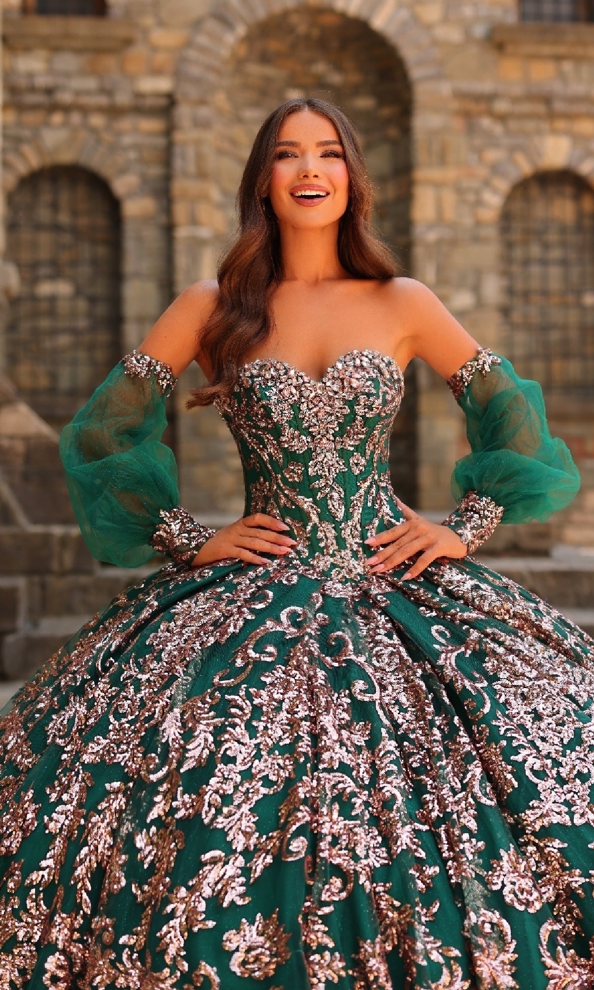 A woman in the Amarra 54238 Sequin Quinceanera Dress, featuring a green ball gown silhouette with a corset bodice, silver embellishments, and sheer sleeves, smiles in front of a stone building.